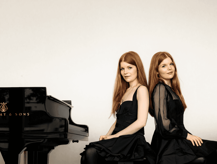 Two women with long red hair sit back-to-back in black dresses next to a grand piano in a minimalist setting.