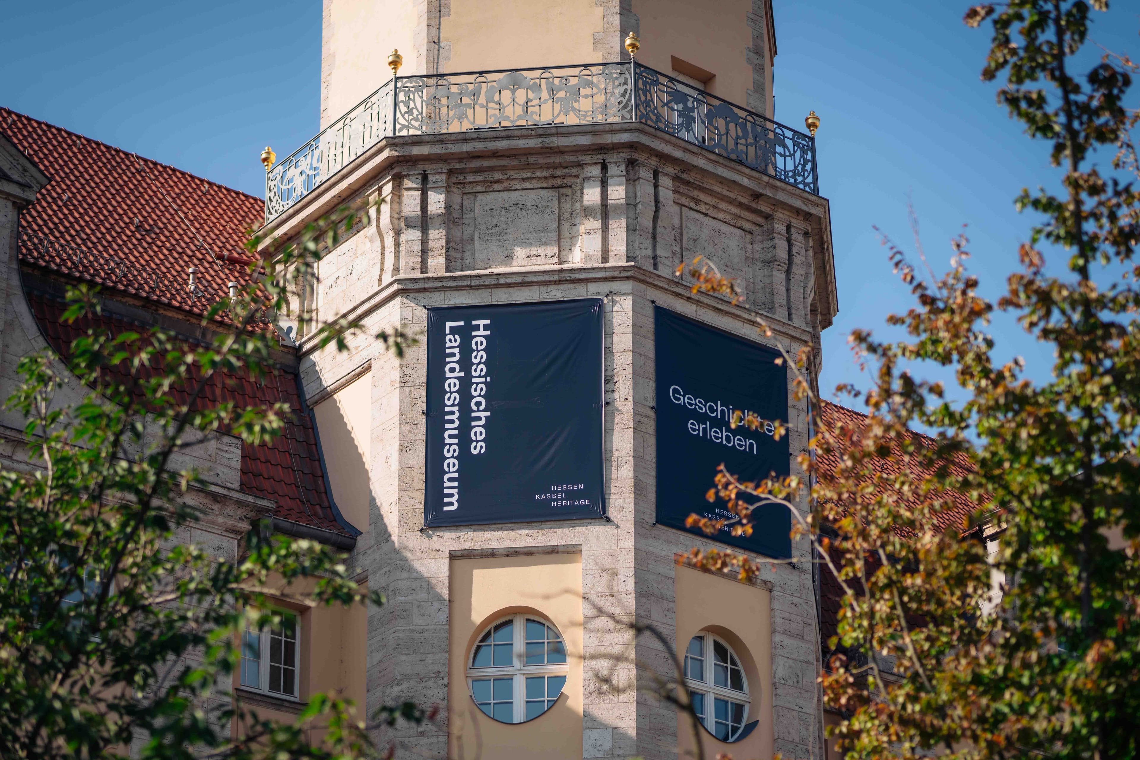 Historic building mit banners für "Hessisches Landesmuseum" und "Geschichte erleben," surrounded by trees unter a clear blue sky.