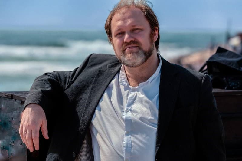 Man with a beard in a black blazer and white shirt leans on a railing by the sea, with waves in the background under a clear blue sky.