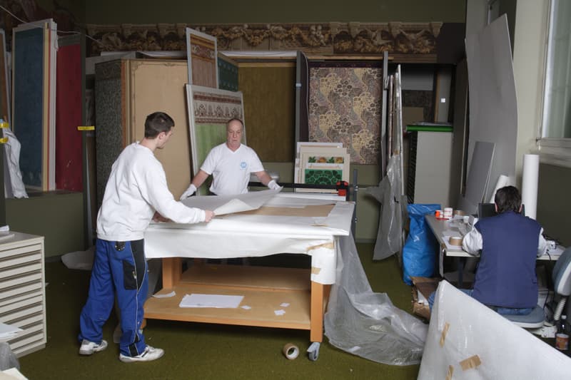 Zwei Männer arbeiten an einem großen Tisch mit Tapetenmustern, während eine andere Person an einem Schreibtisch in einem überfüllten Raum sitzt.