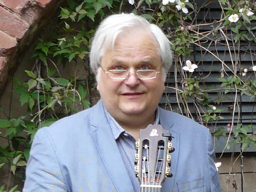 Man with white hair and glasses, wearing a blue jacket, holding a guitar. Background features a brick arch and flowering vines.