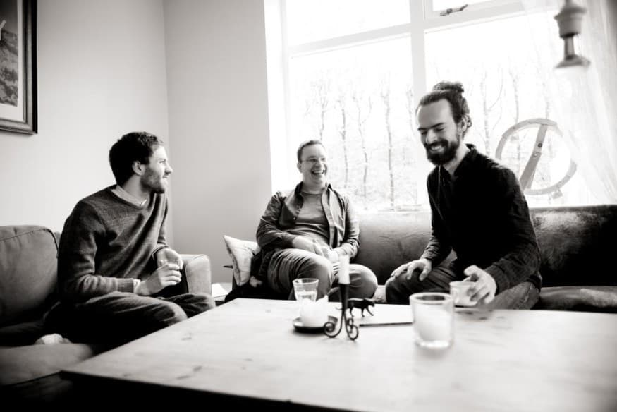 Three men sitting and laughing around a coffee table in a cozy living room with a large window in the background. Black and white photo.