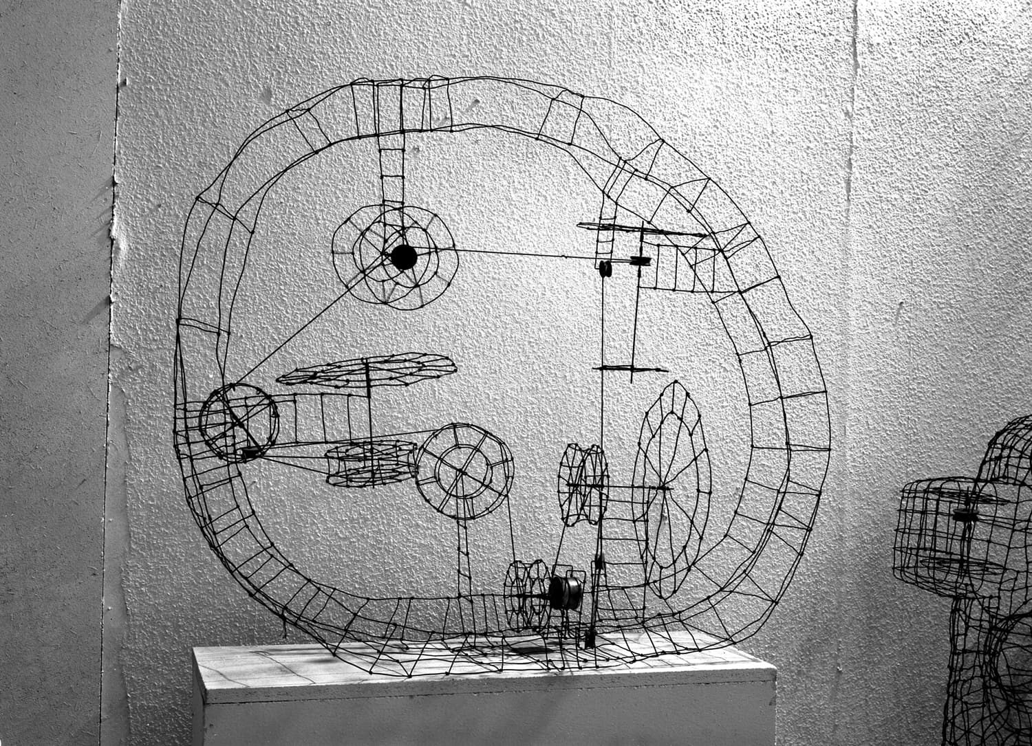 Drahtskulptur, die einer komplexen mechanischen Struktur mit kreisförmigen und linearen Elementen ähnelt und auf einem weißen Sockel vor einer strukturierten Wand ausgestellt ist.