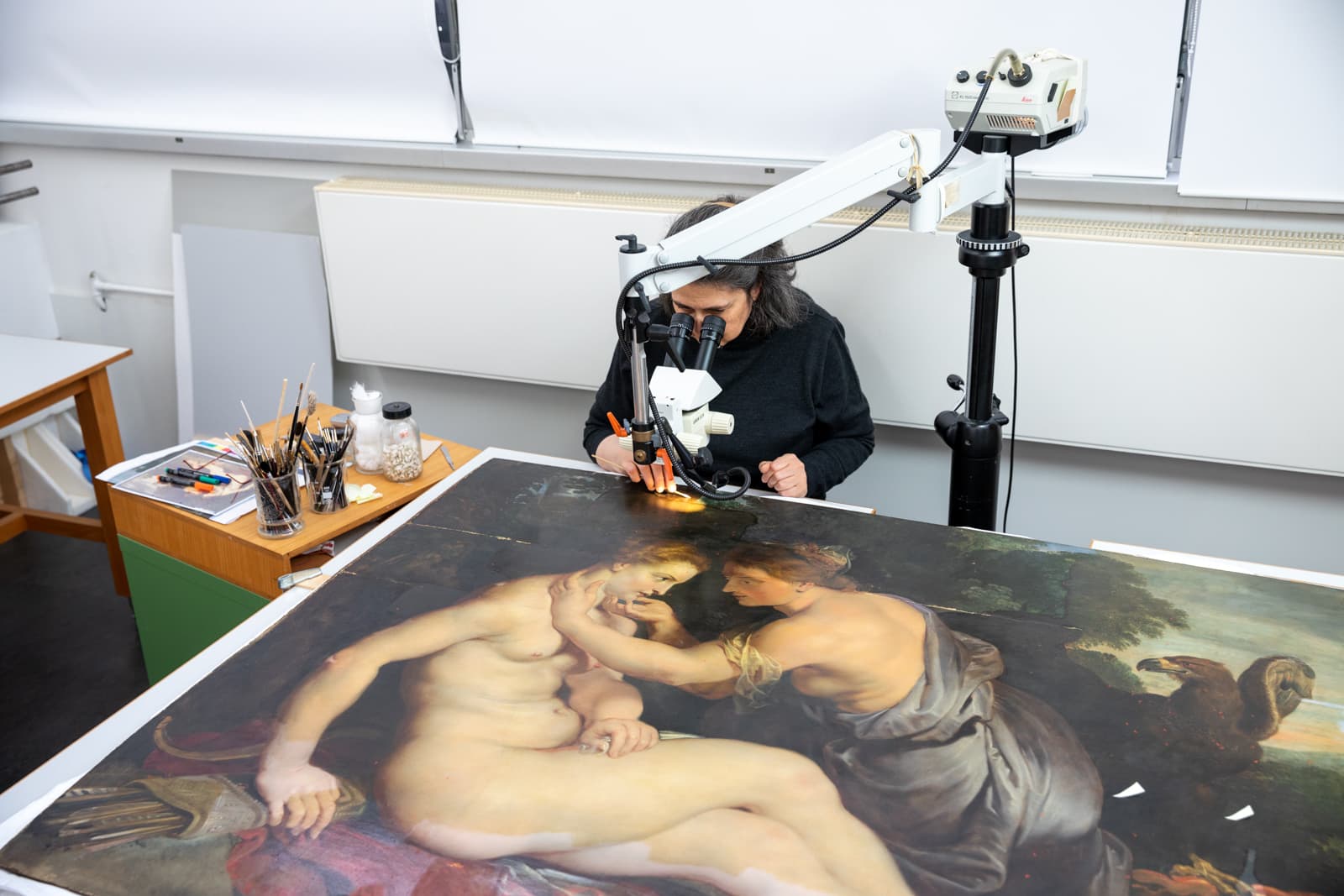 A conservator examines a large painting with a microscope, surrounded by brushes and tools on a table in an art restoration studio.