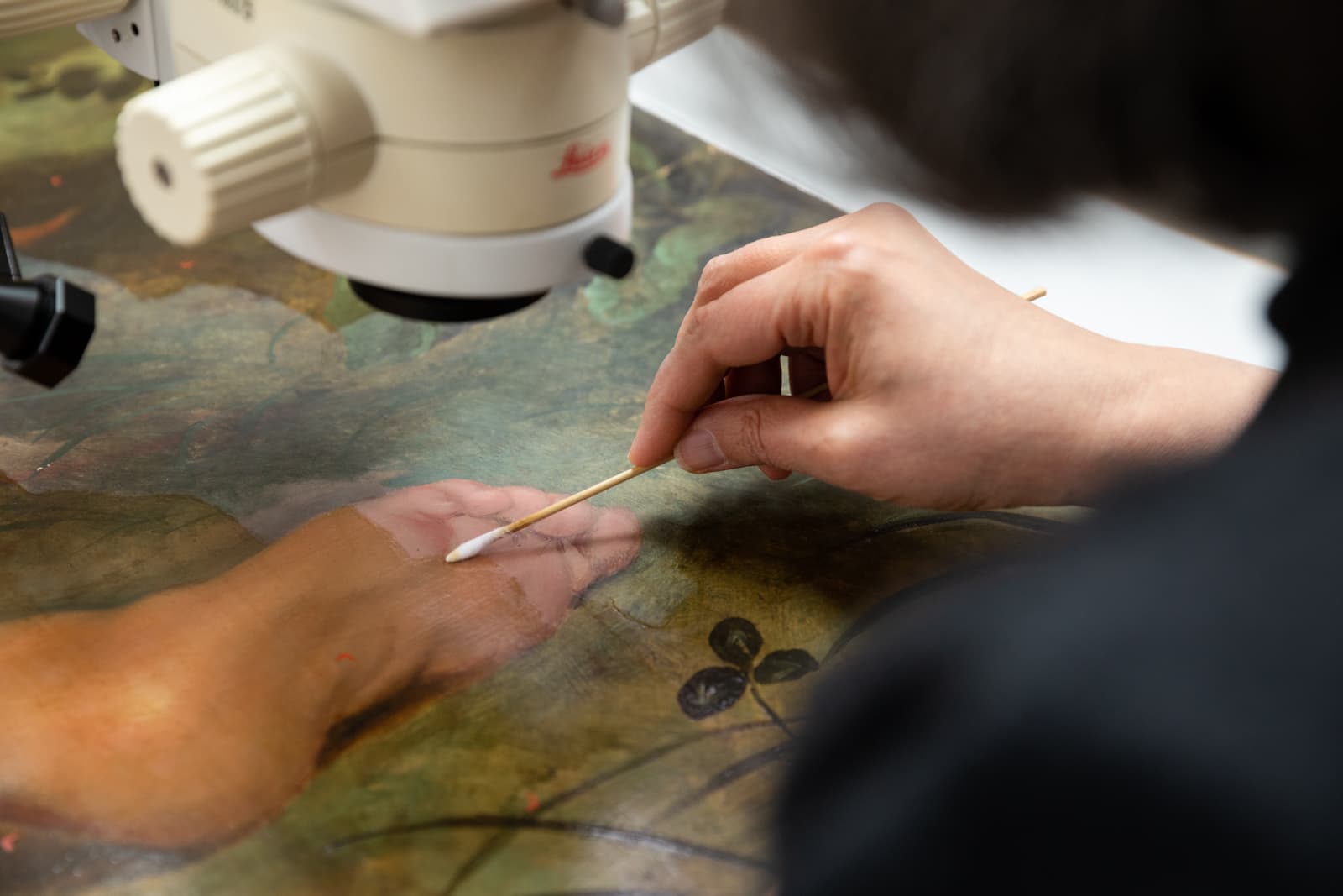 A person uses a swab and magnifier to delicately restore a painting, focusing on details of a hand and floral elements.