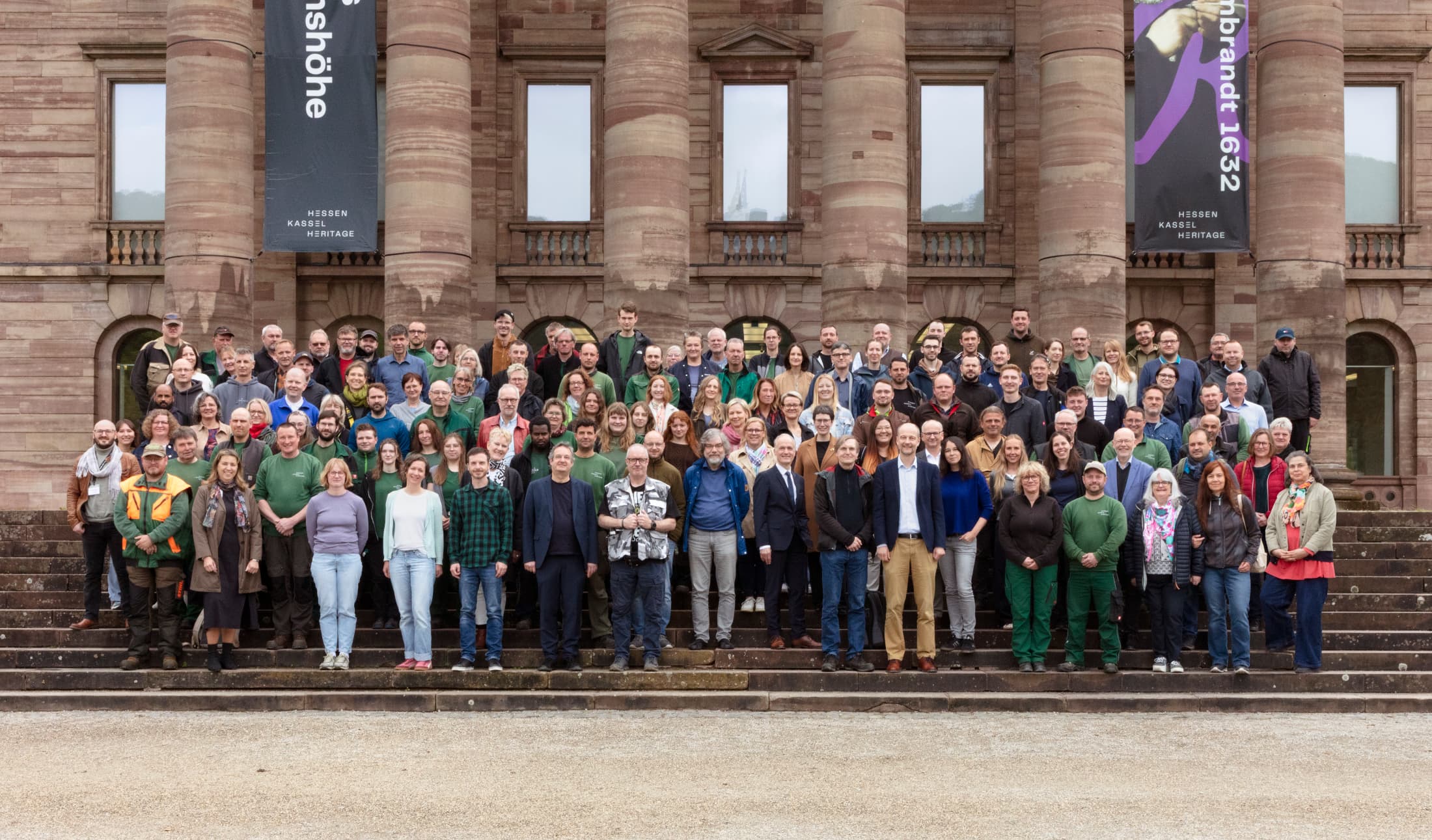 Eine große Gruppe von Menschen posiert auf den Stufen vor einem historischen Gebäude mit Säulen und Fahnen.