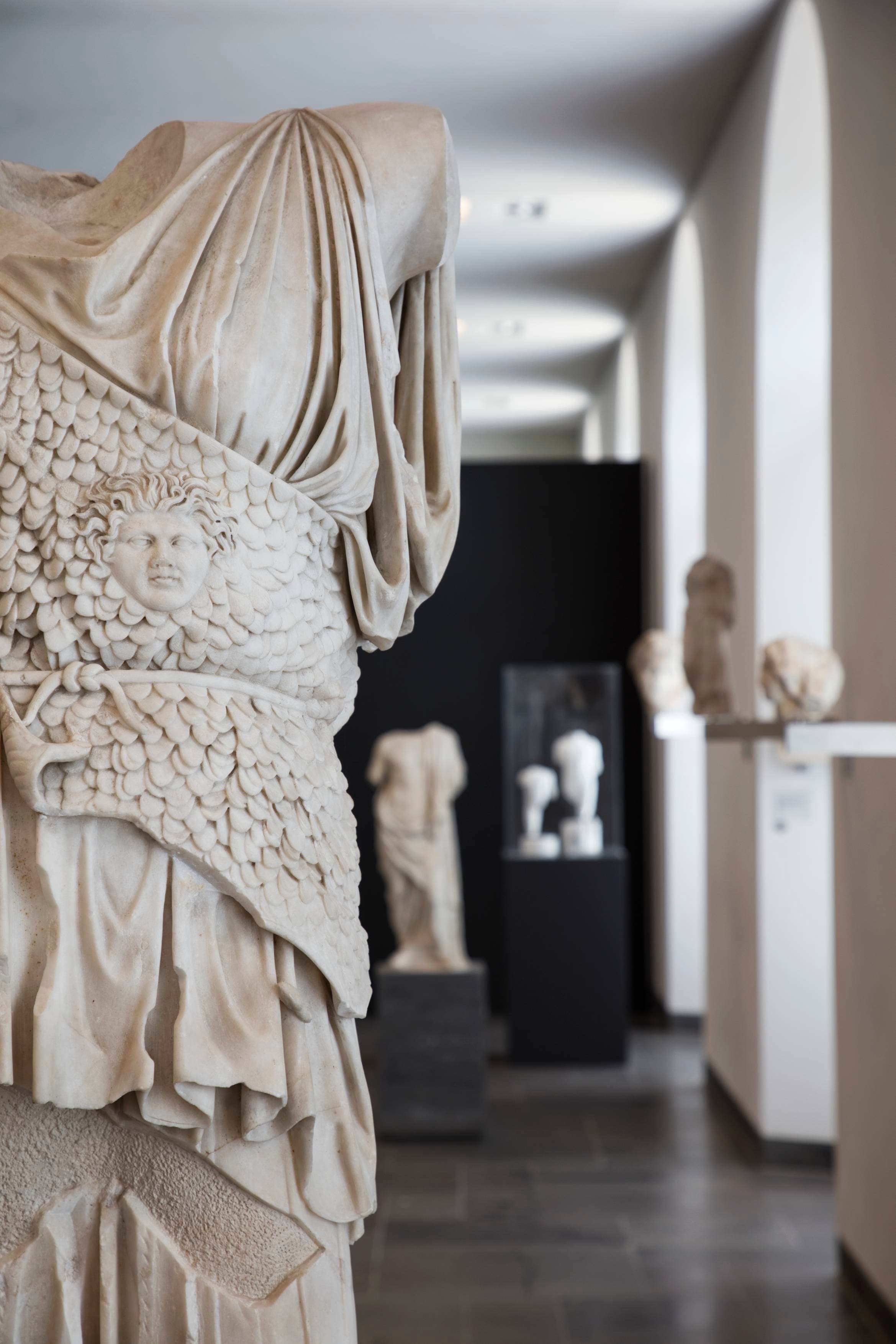 Marble statue of a torso with intricate armor in a museum gallery, surrounded by other classical sculptures on display.