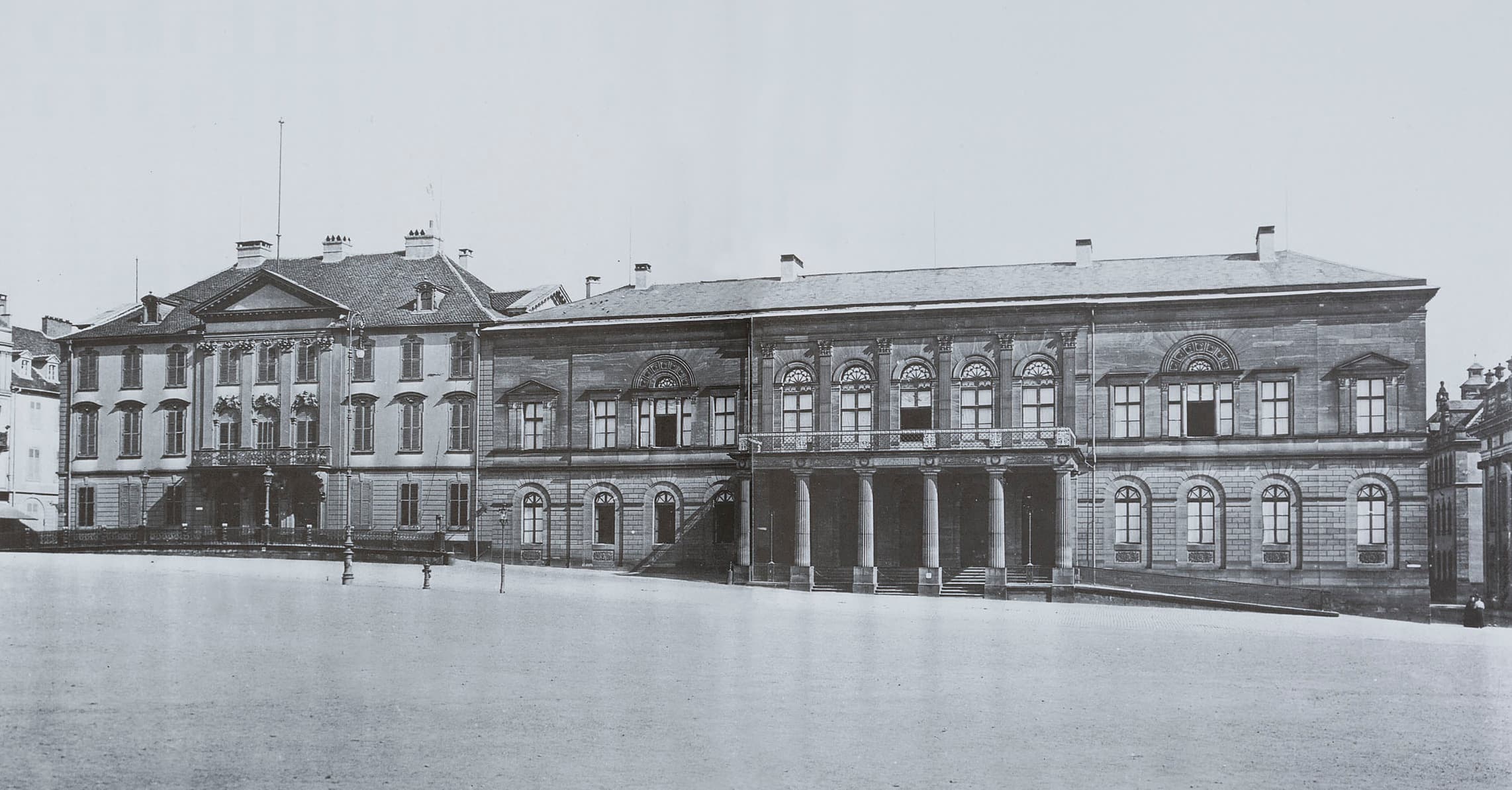 Historisches Schwarzweißfoto eines stattlichen zweiflügeligen Gebäudes mit großen Fenstern, Säulen und einem geräumigen Innenhof davor.