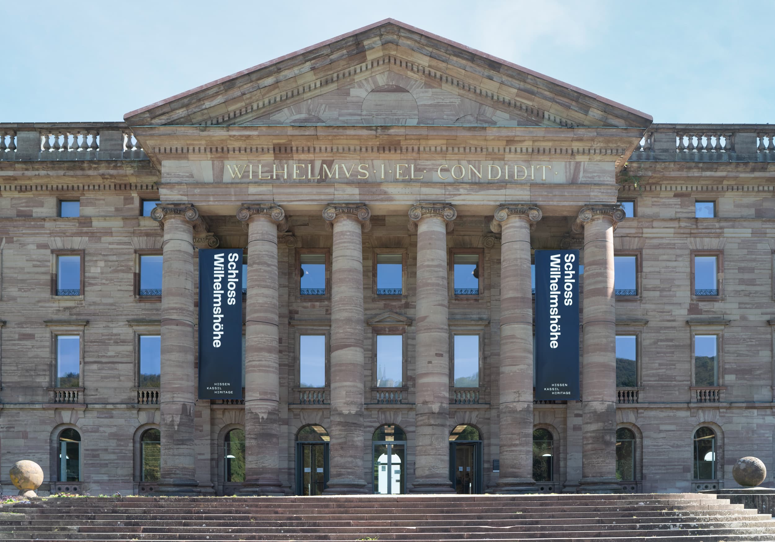 Front view of a grand neoclassical building with six large columns, banners, and wide steps leading to the entrance.