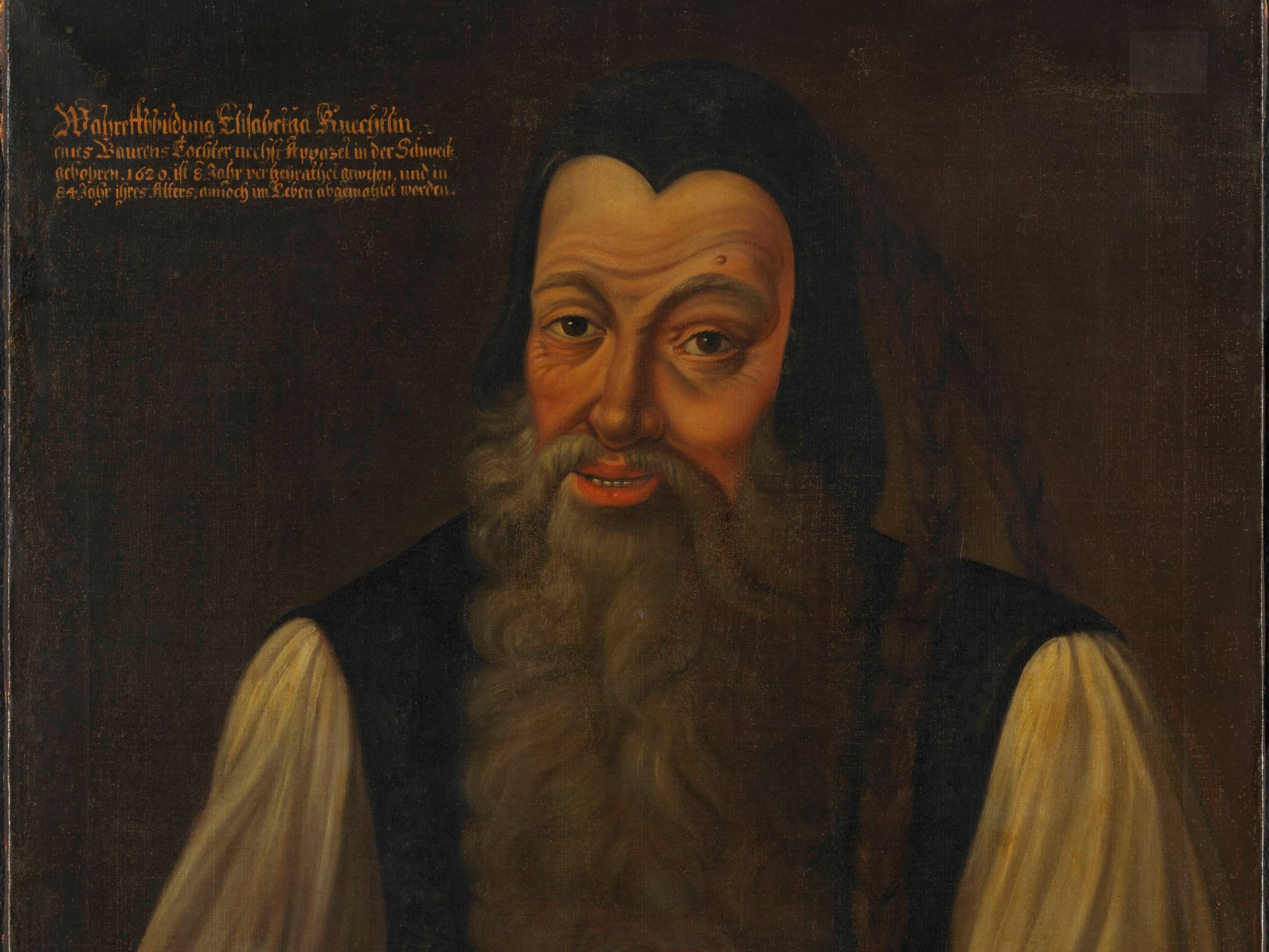 Portrait of a bearded man wearing a traditional outfit with a red vest and white shirt, set against a dark background.