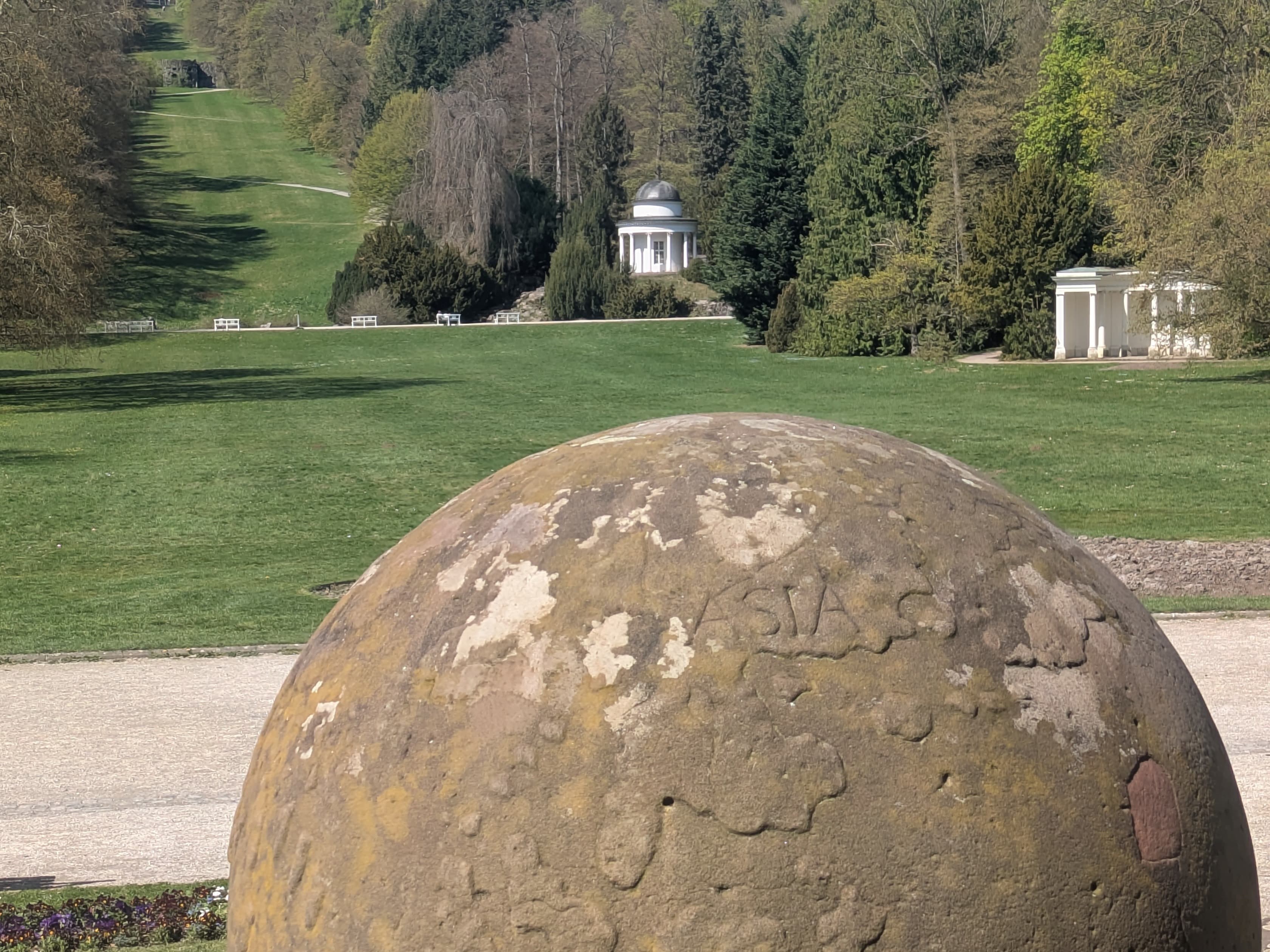 Stone sphere in foreground, with a long garden path leading to a distant palace and pavilion, surrounded by trees and greenery.