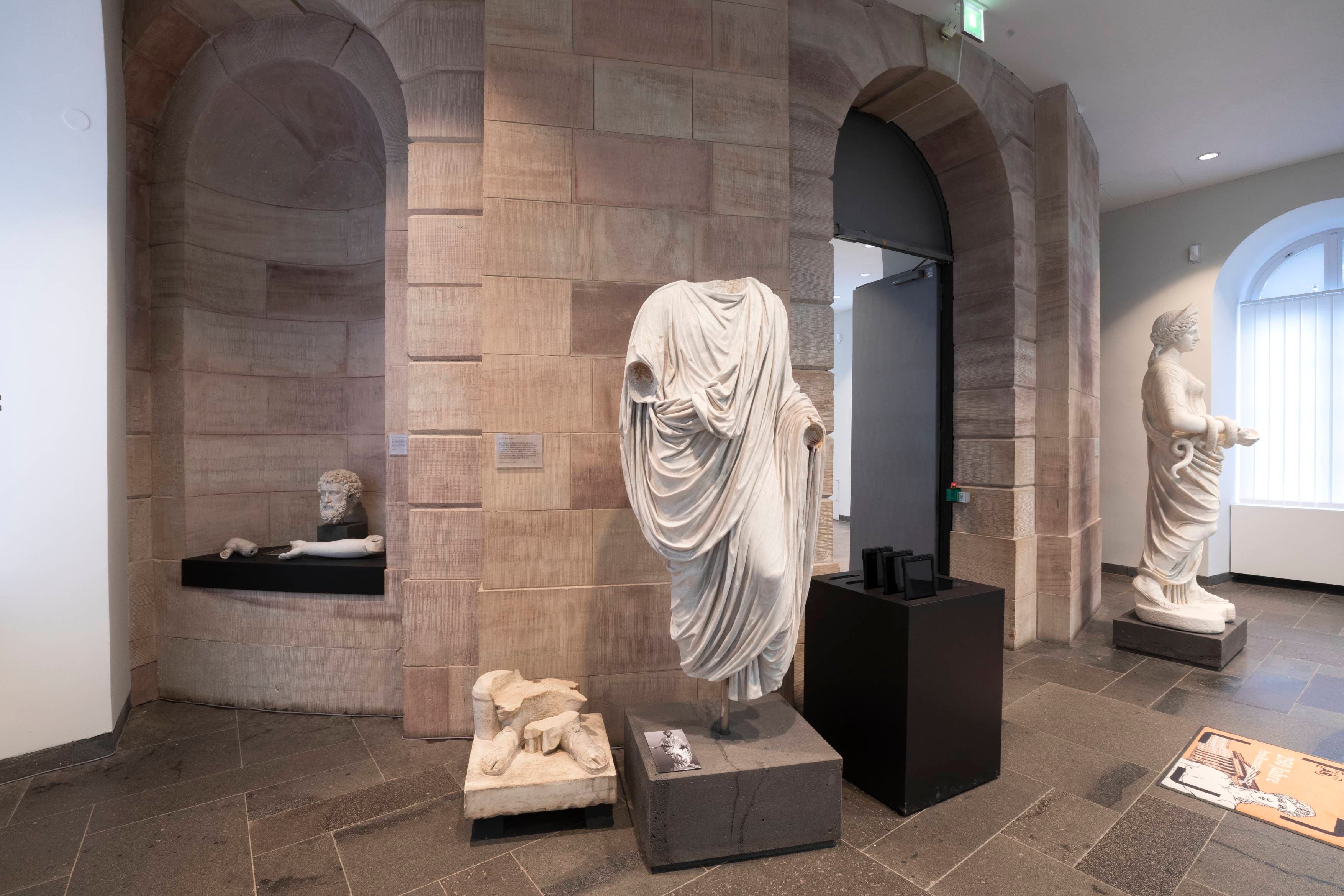Museumsausstellung mit klassischen Skulpturen, darunter eine drapierte Figur, eine liegende Statue und eine stehende Figur, vor Steinmauern.