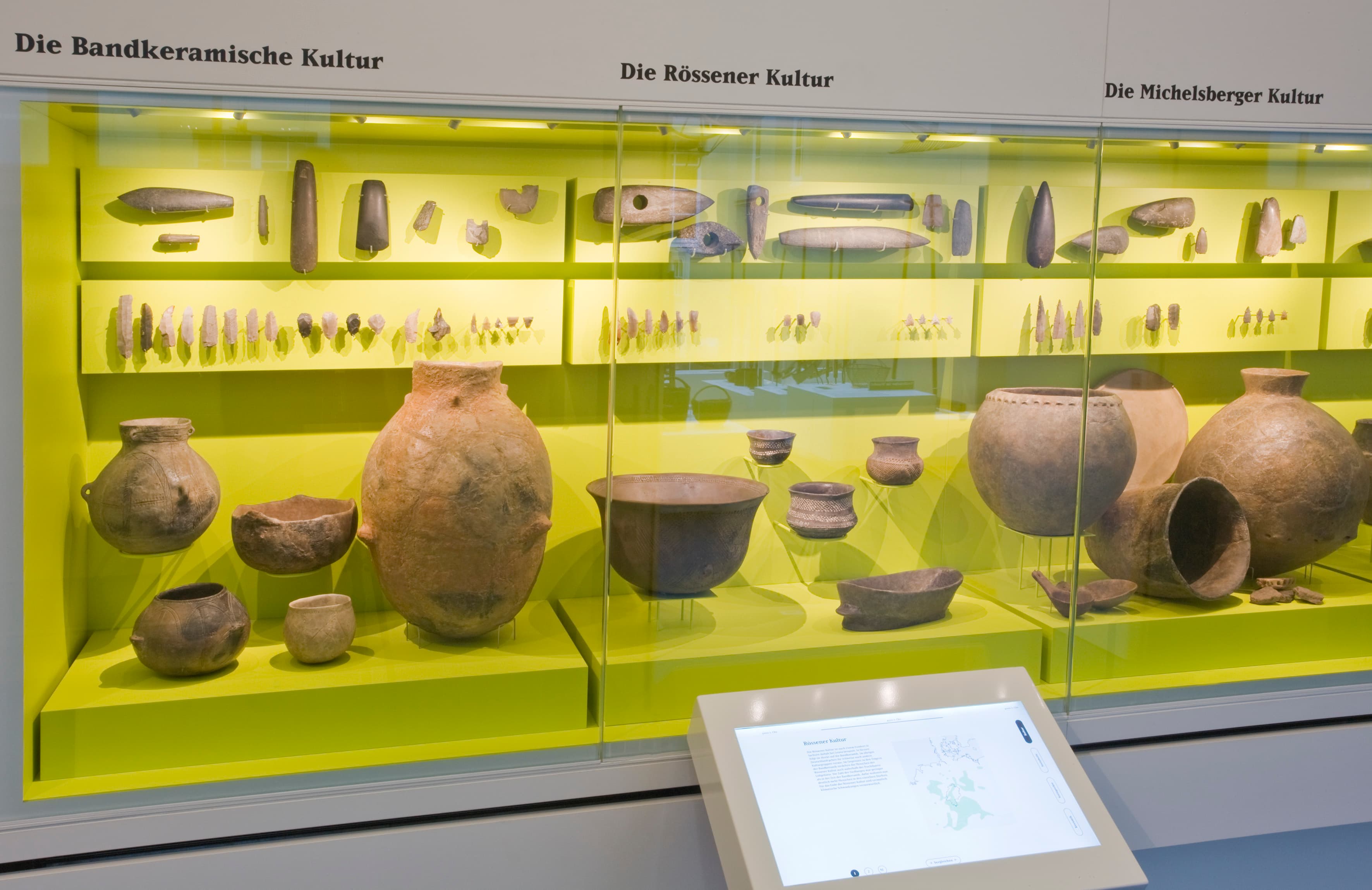 Ausstellung antiker Töpferwaren und Werkzeuge in einer Museumsausstellung mit gelber Hintergrundbeleuchtung und Informationstafeln darüber.