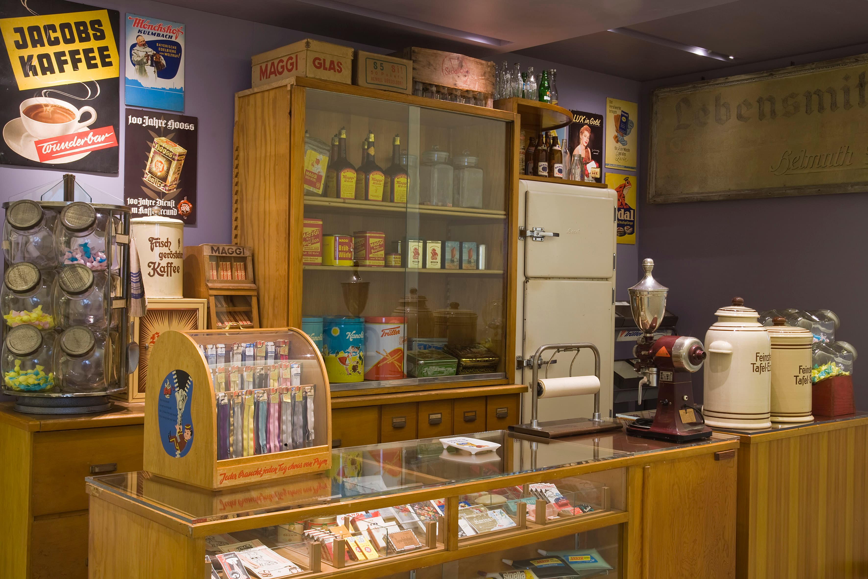 Vintage-Ladeneinrichtung mit Holzregalen, Retro-Werbung, einer Glasvitrine mit verschiedenen Waren und altmodischen Küchengeräten.
