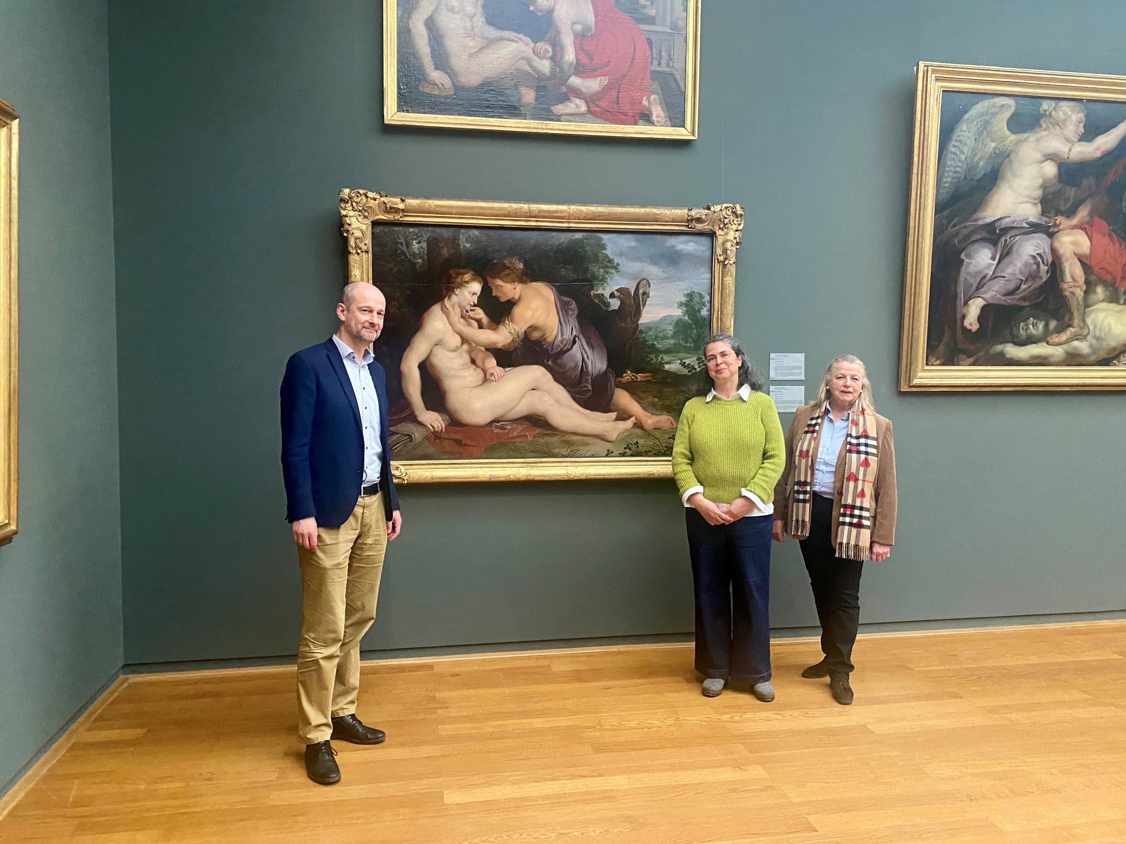 Three people stand in an art gallery, posing in front of a large framed painting. The room has wooden floors and blue-green walls.