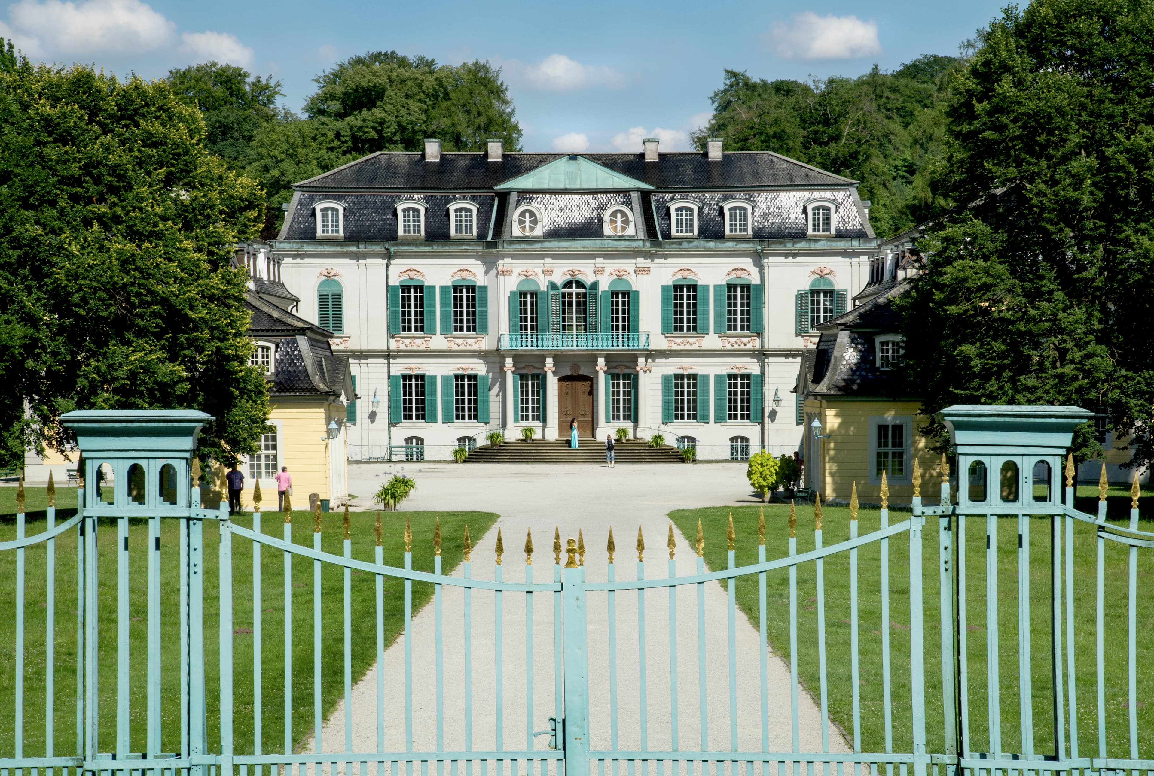 Elegantes europäisches Herrenhaus mit blaugrünen Fensterläden und verzierter Fassade, umgeben von üppigem Grün und eingerahmt von einem offenen schmiedeeisernen Tor.