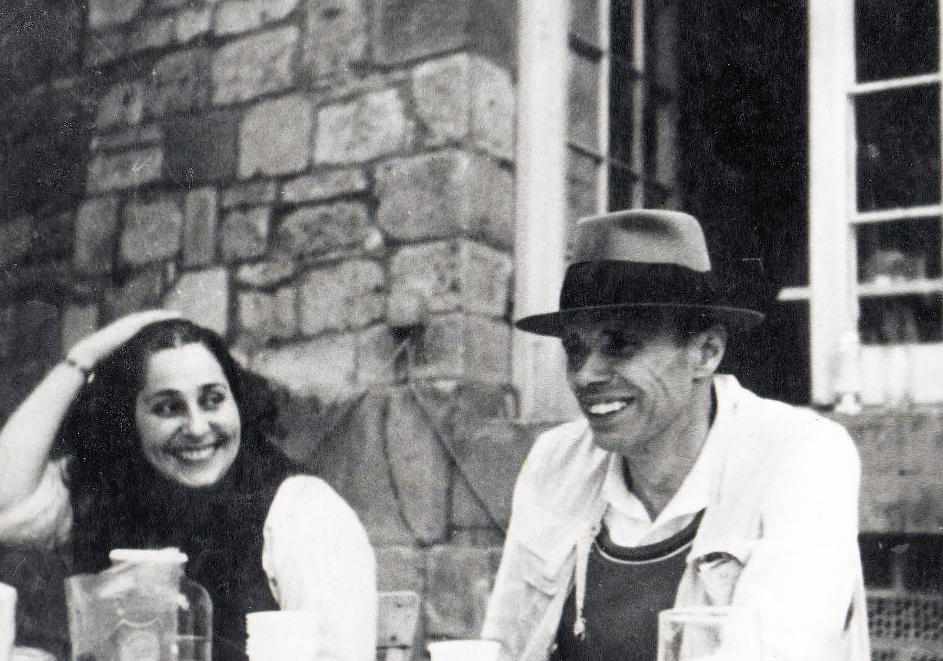 A woman and a man in a hat sit outside by a stone building, smiling and enjoying a casual moment together.