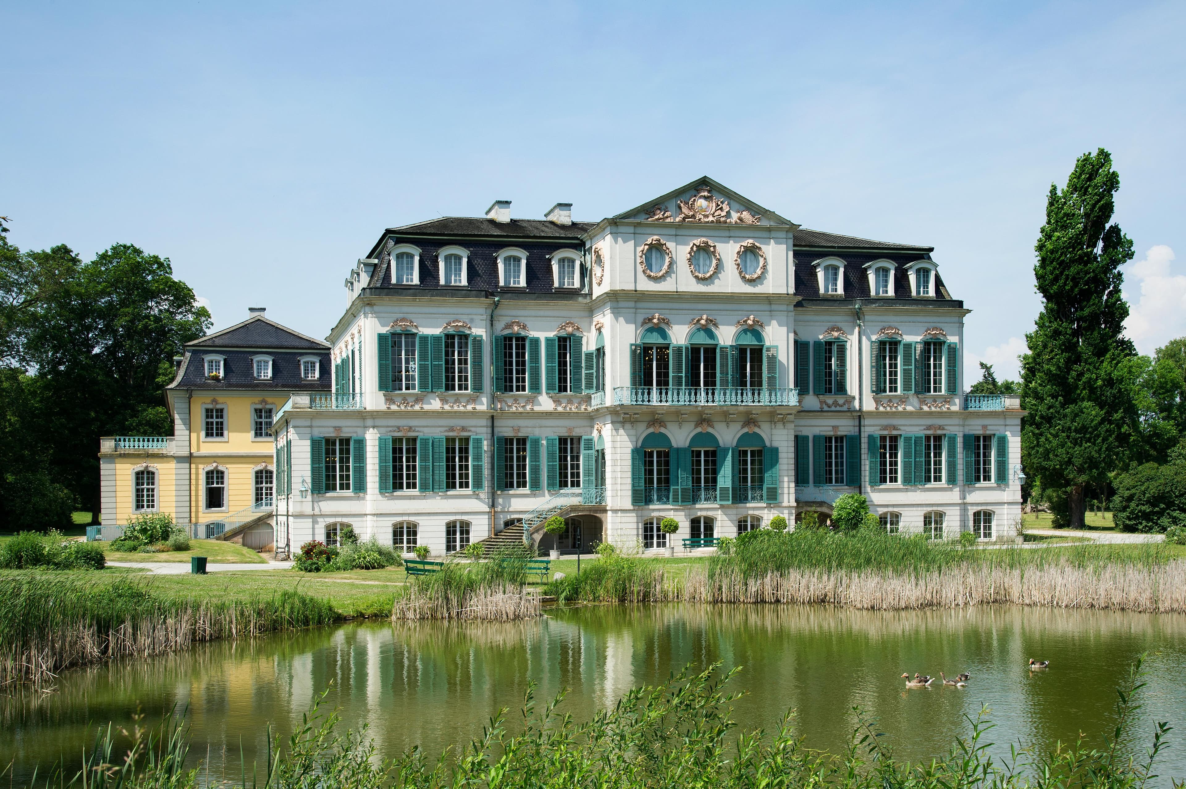 Ein großes, historisches Herrenhaus mit grünen Fensterläden und kunstvollen Details liegt an einem ruhigen Teich, umgeben von üppigem Grün unter einem klaren blauen Himmel.