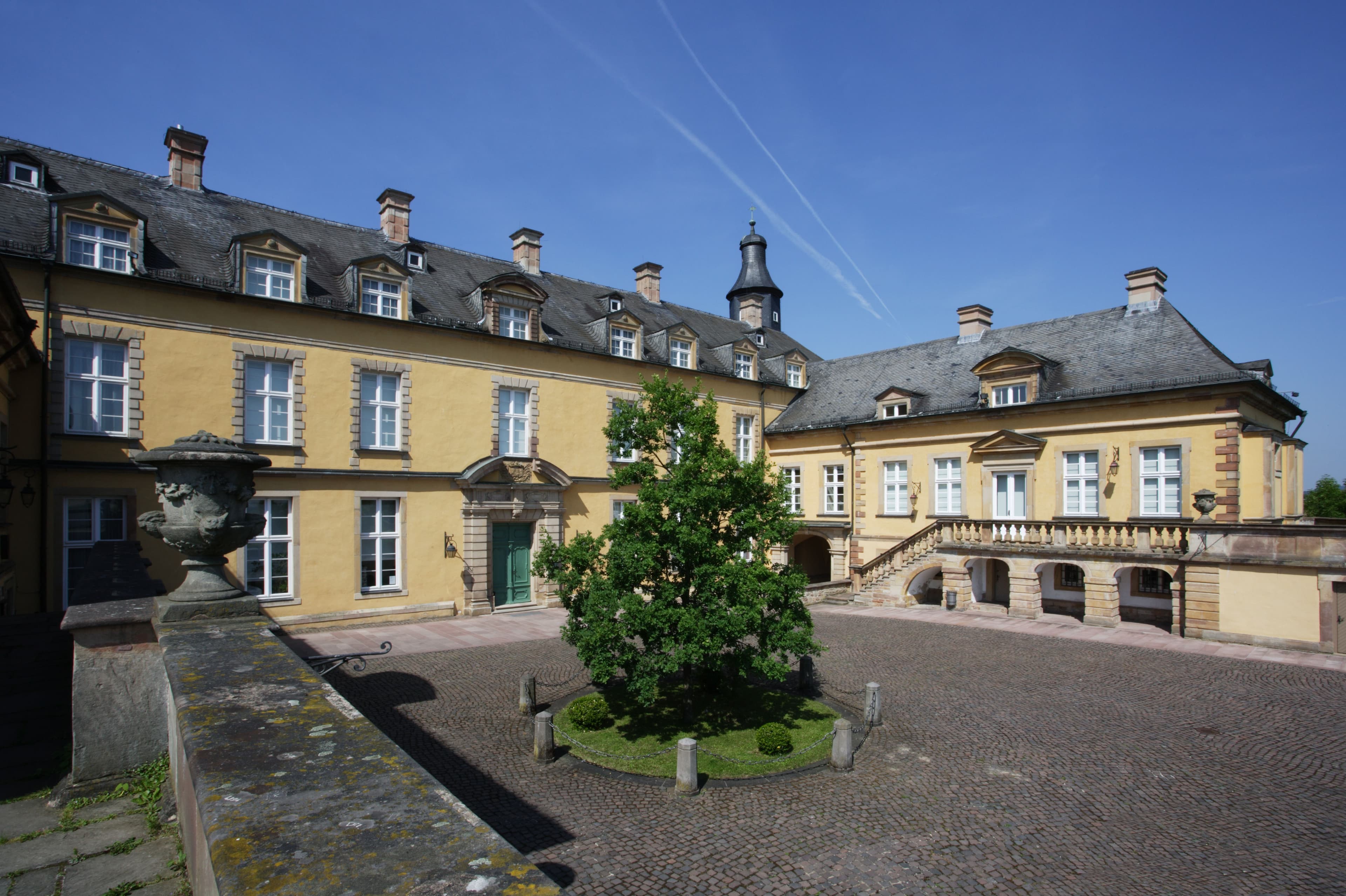 Historisches gelbes Gebäude mit Schieferdach, mehreren Schornsteinen und einem zentralen Innenhof mit einem runden Garten und einem Baum unter einem klaren blauen Himmel.