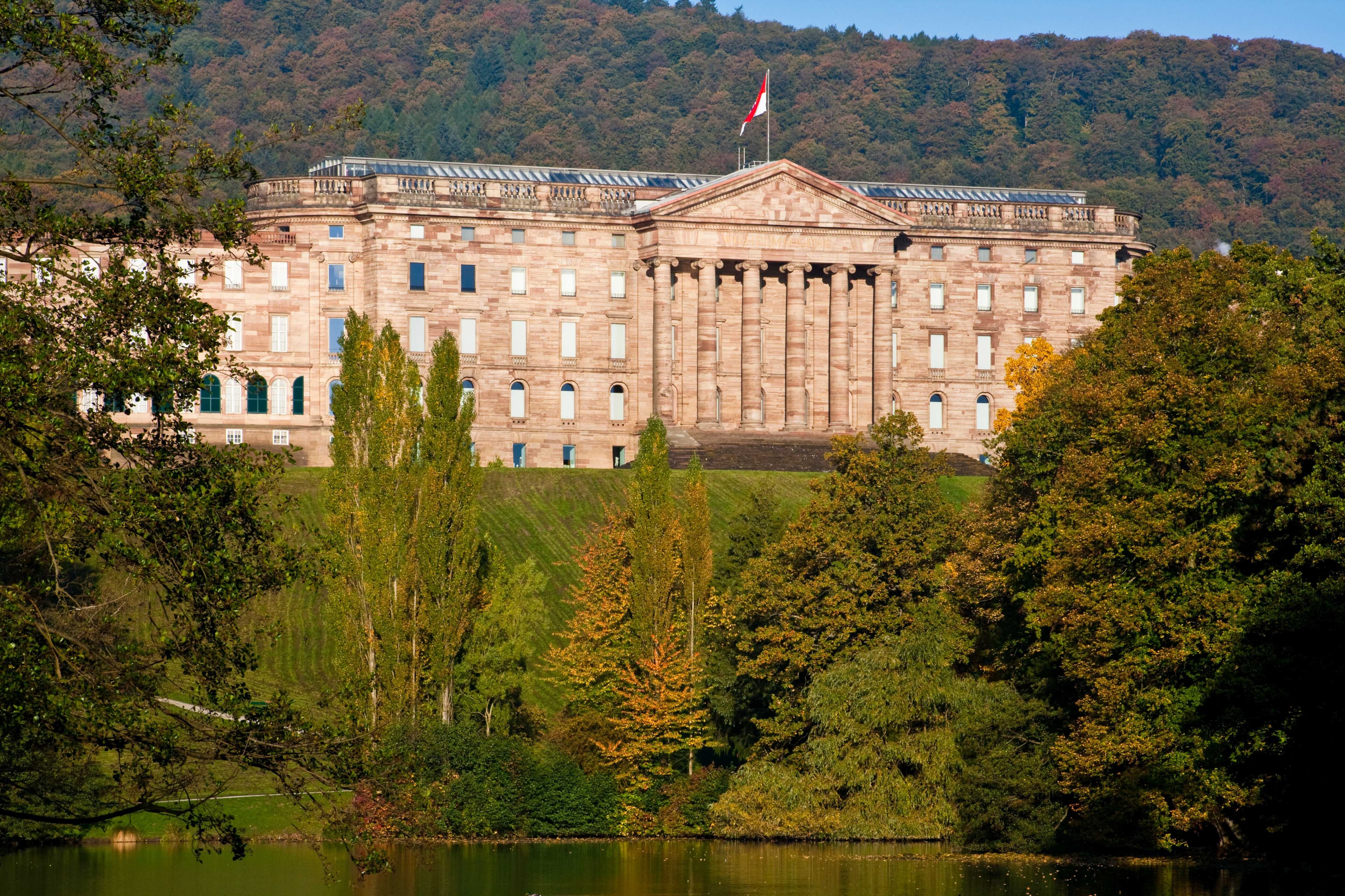 Neoklassizistischer Palast mit Säulen und einer Flagge auf der Spitze, umgeben von Bäumen und einem See, vor der Kulisse eines bewaldeten Hügels.