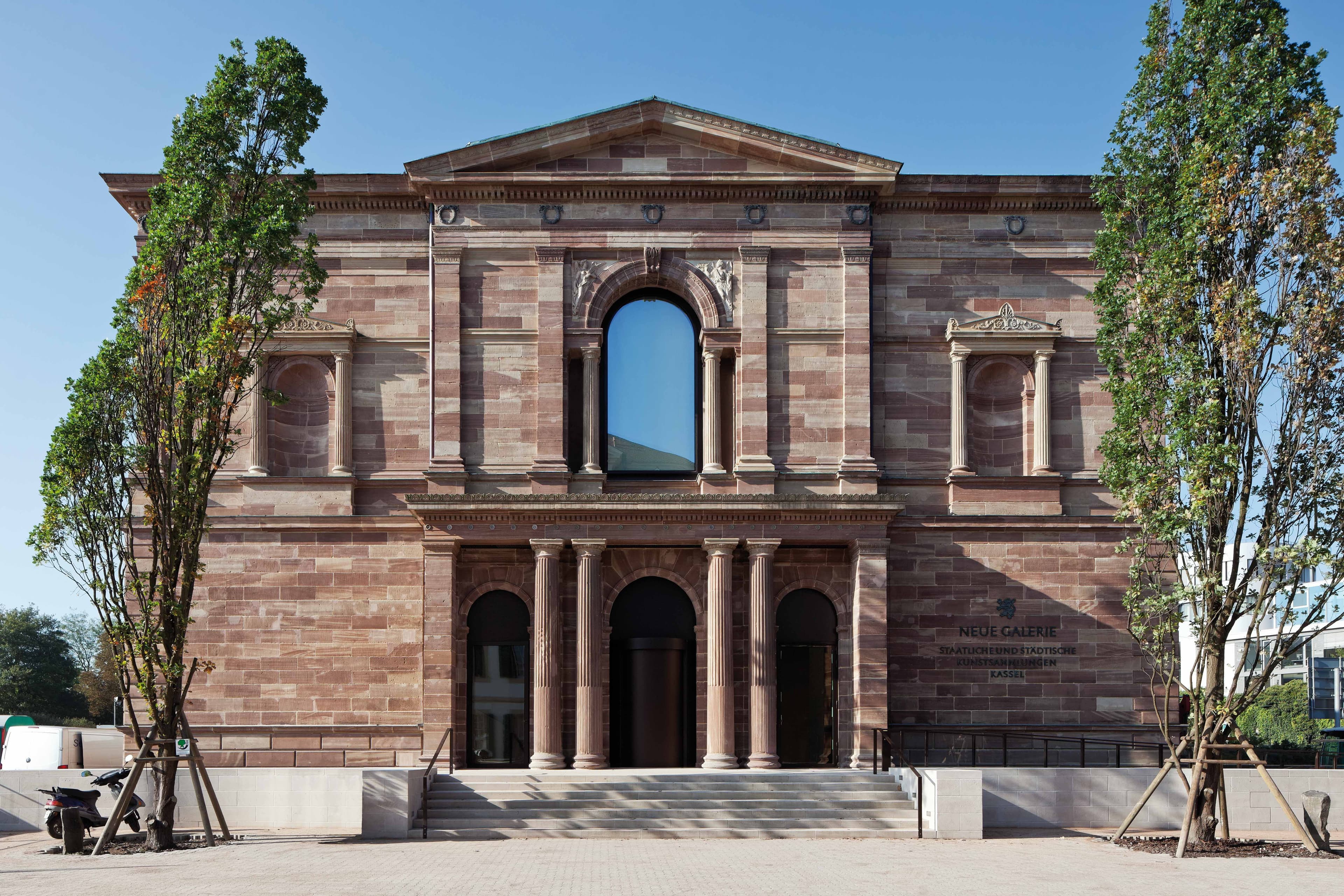 Historic stone building with tall arched windows and columns, flanked by trees, under a clear blue sky.