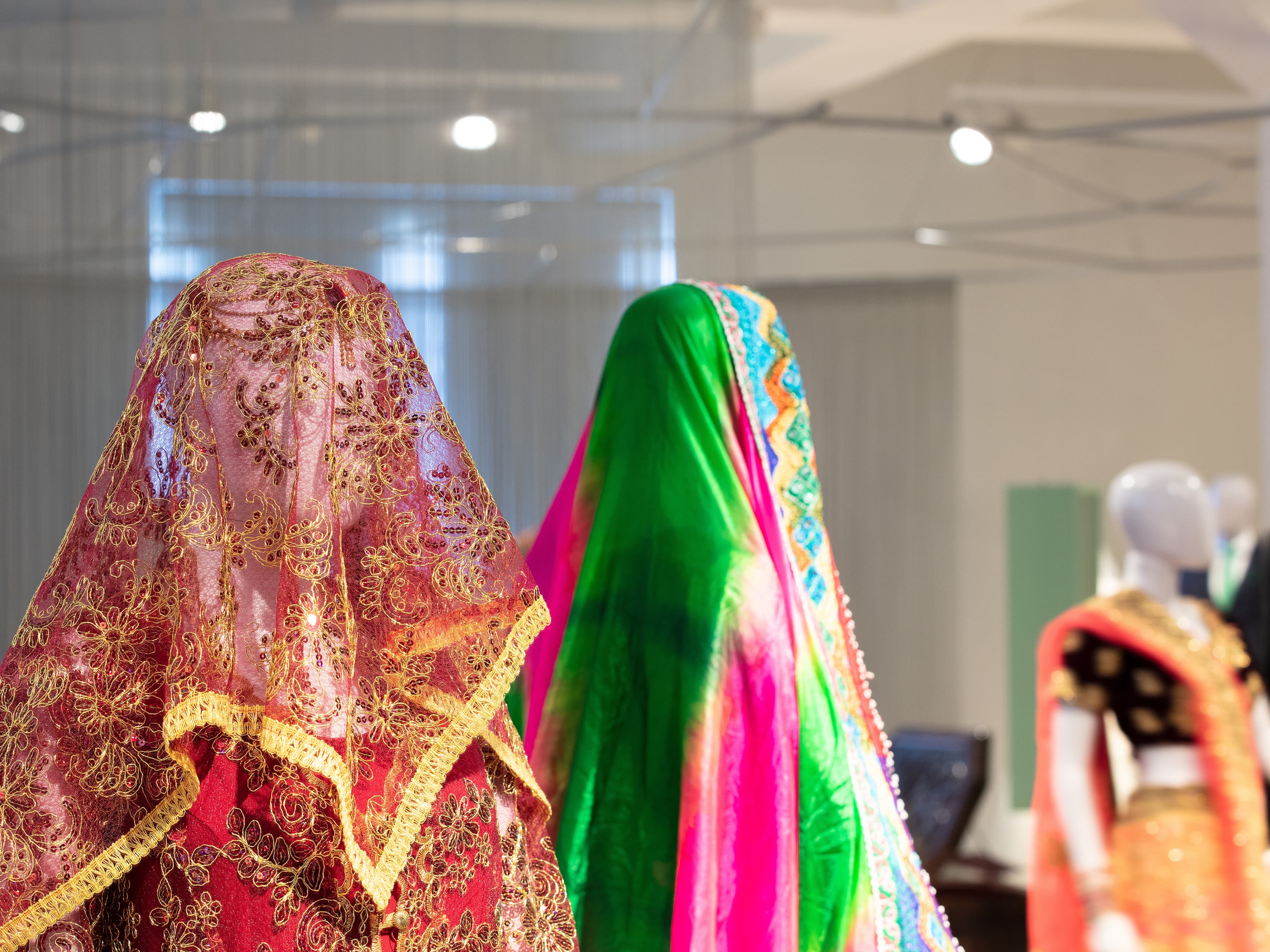 Two mannequins draped in vibrant, ornate fabrics; one in red and gold, the other in green and pink, displayed in a well-lit showroom.