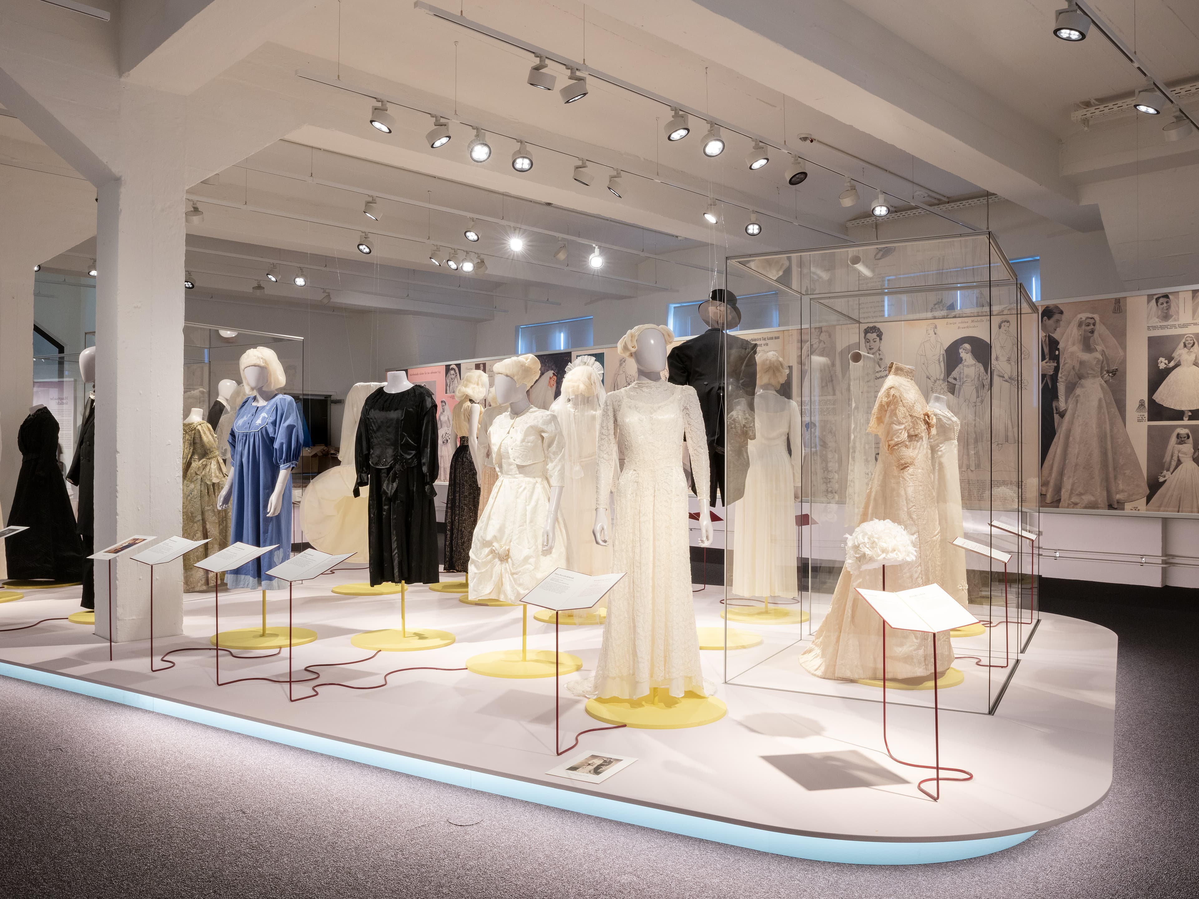 Exhibit of vintage wedding dresses displayed on mannequins in a well-lit museum setting, with informational plaques beside each dress.