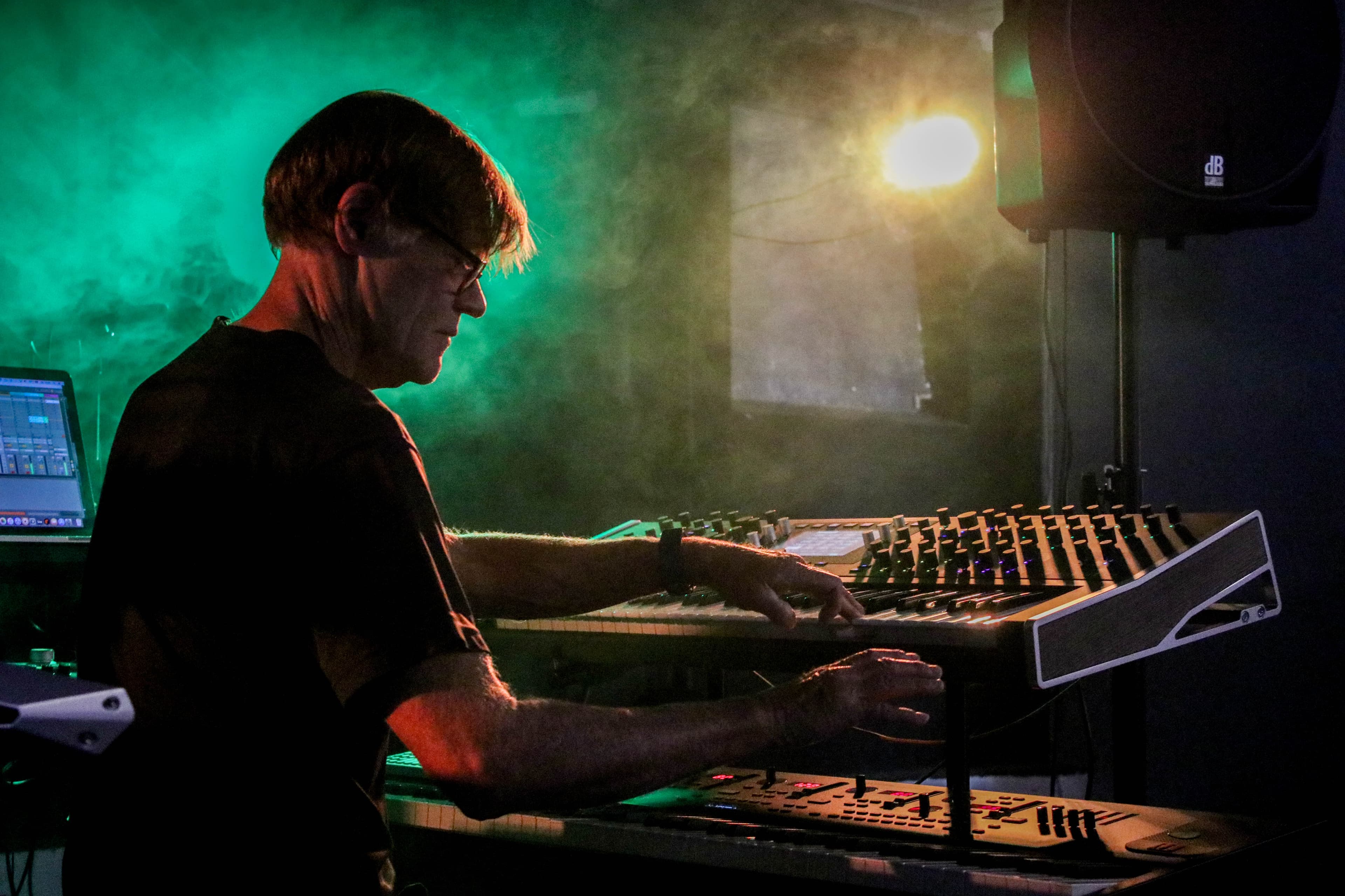 A musician playing keyboards and synthesizers on stage, surrounded by green lighting and smoke.