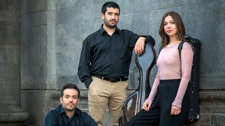 Three people posing outdoors; one is seated, and two are standing near a cello case against a stone wall.