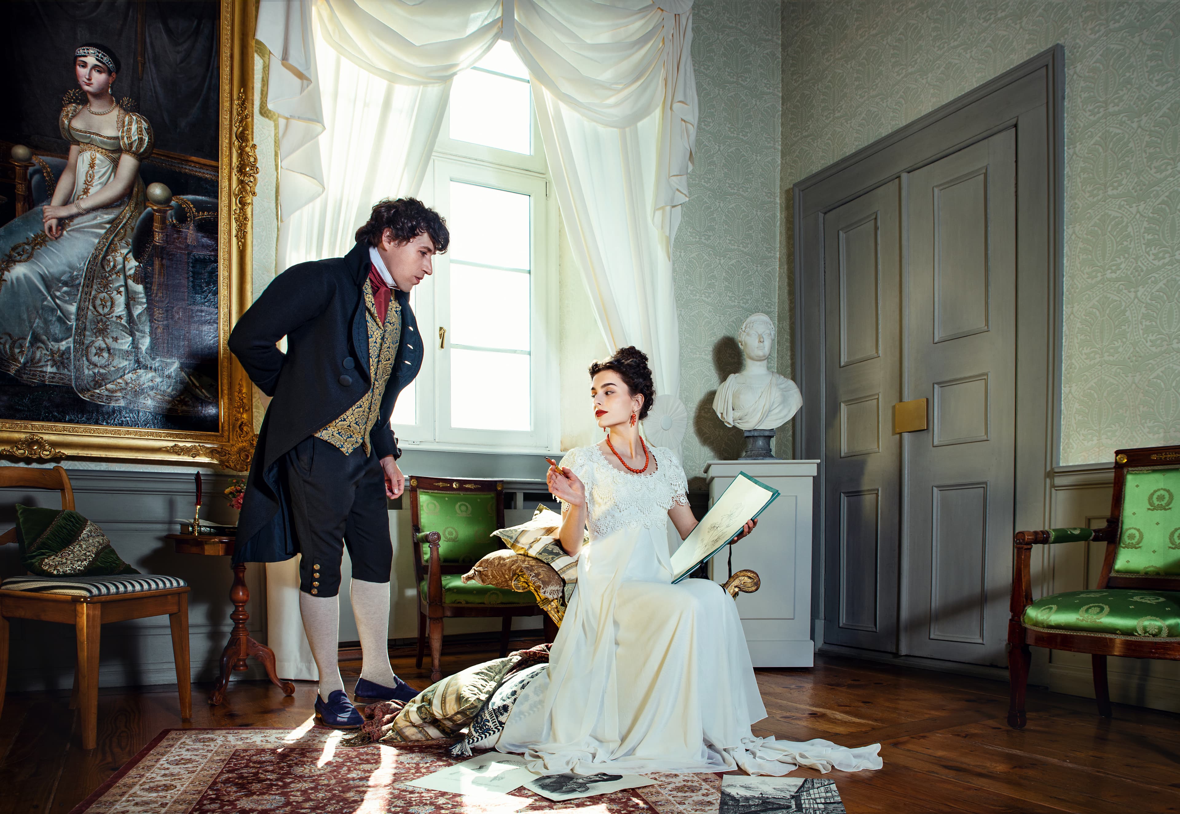 A man in period clothing stands beside a seated woman in a white dress holding a book, in an elegant room with a large painting and a bust.