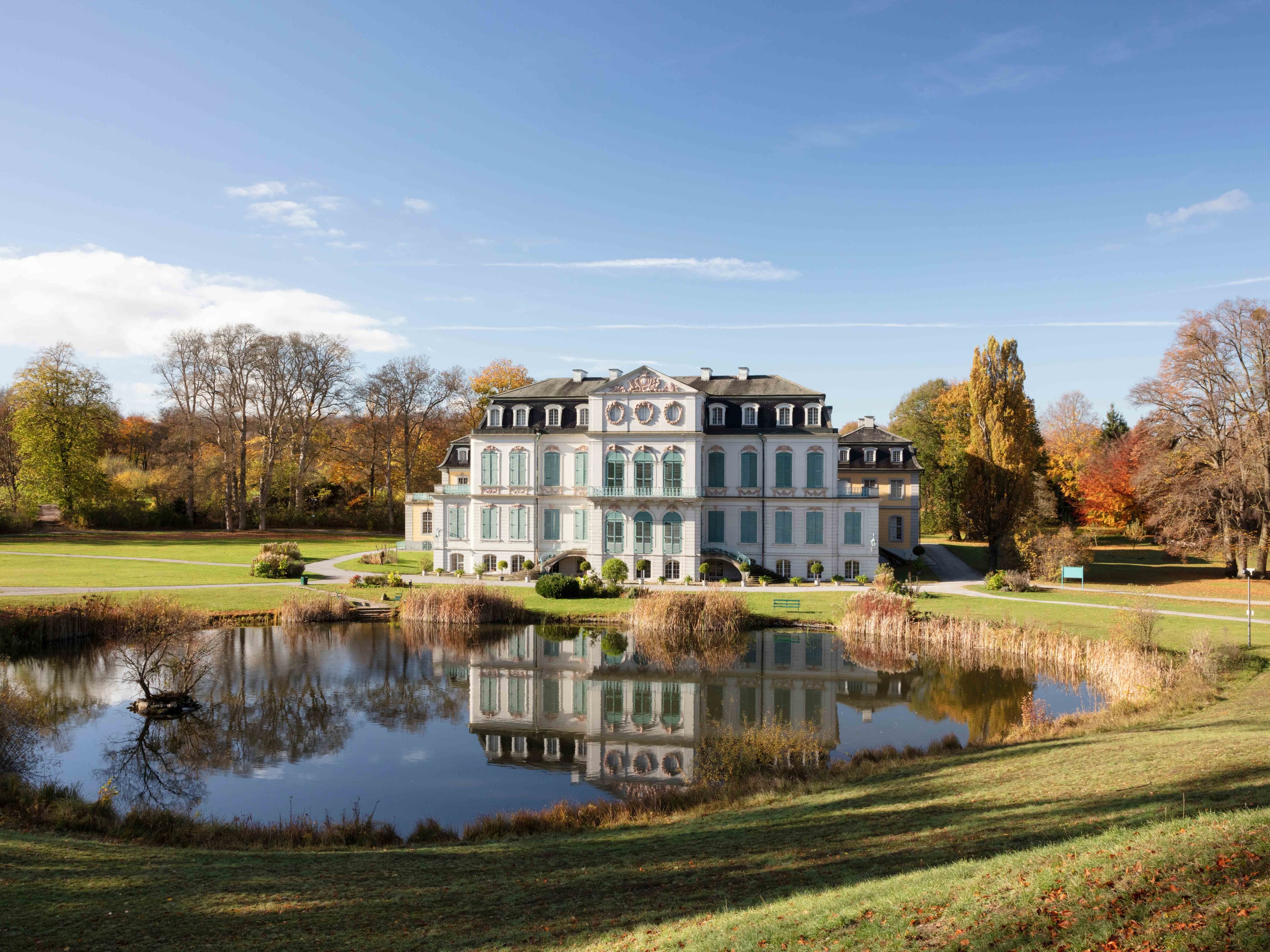 Elegantes Herrenhaus mit blauen Fensterläden und kunstvollen Details, das sich in einem ruhigen Teich spiegelt, umgeben von Herbstbäumen und einem klaren blauen Himmel.