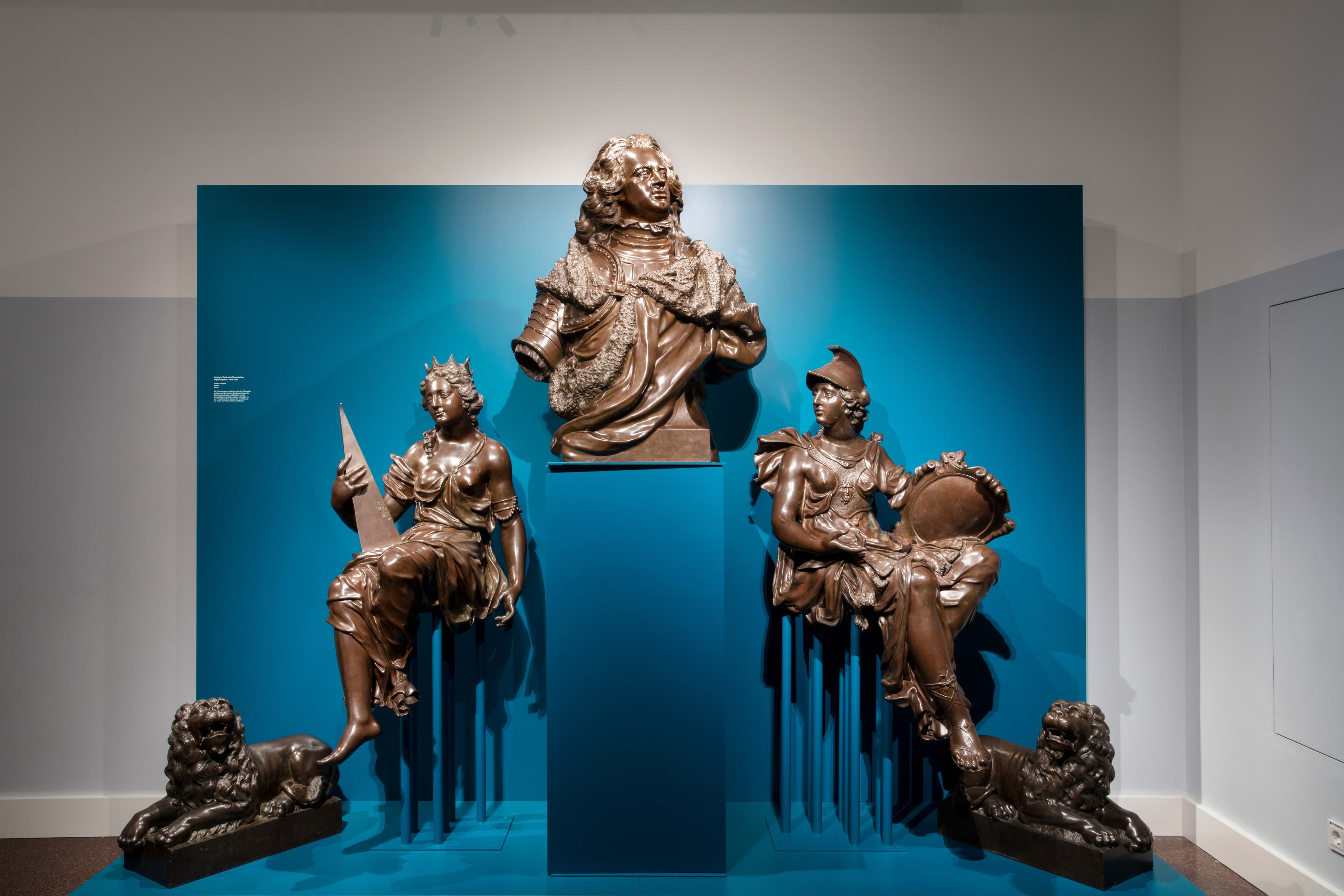 Drei Bronzeskulpturen auf blauem Hintergrund: eine zentrale Büste und zwei sitzende Figuren, jeweils mit einem Löwen zu ihren Füßen.