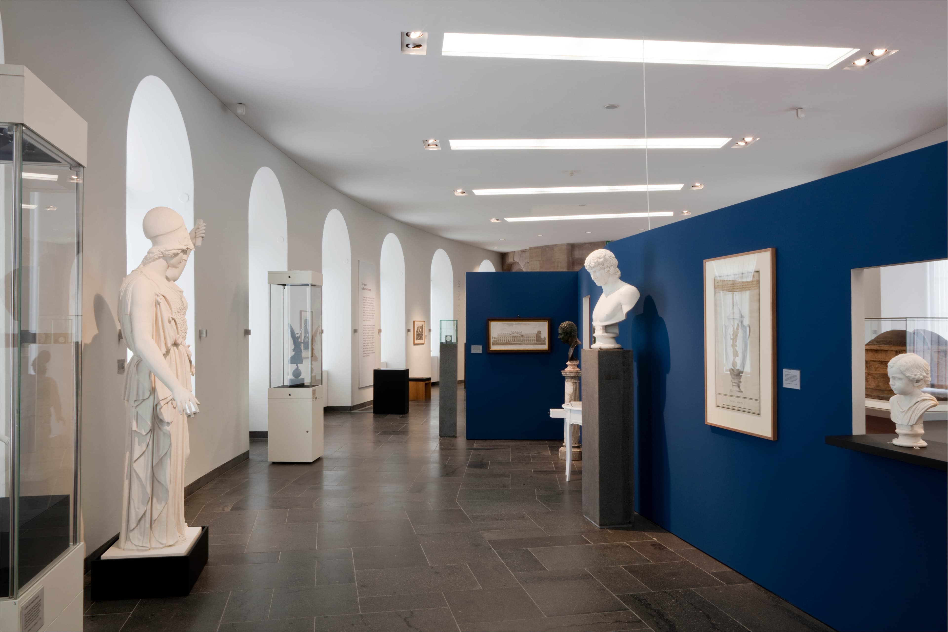 Museum gallery with white sculptures, blue walls, and framed artworks. Arched windows on the left illuminate the space.