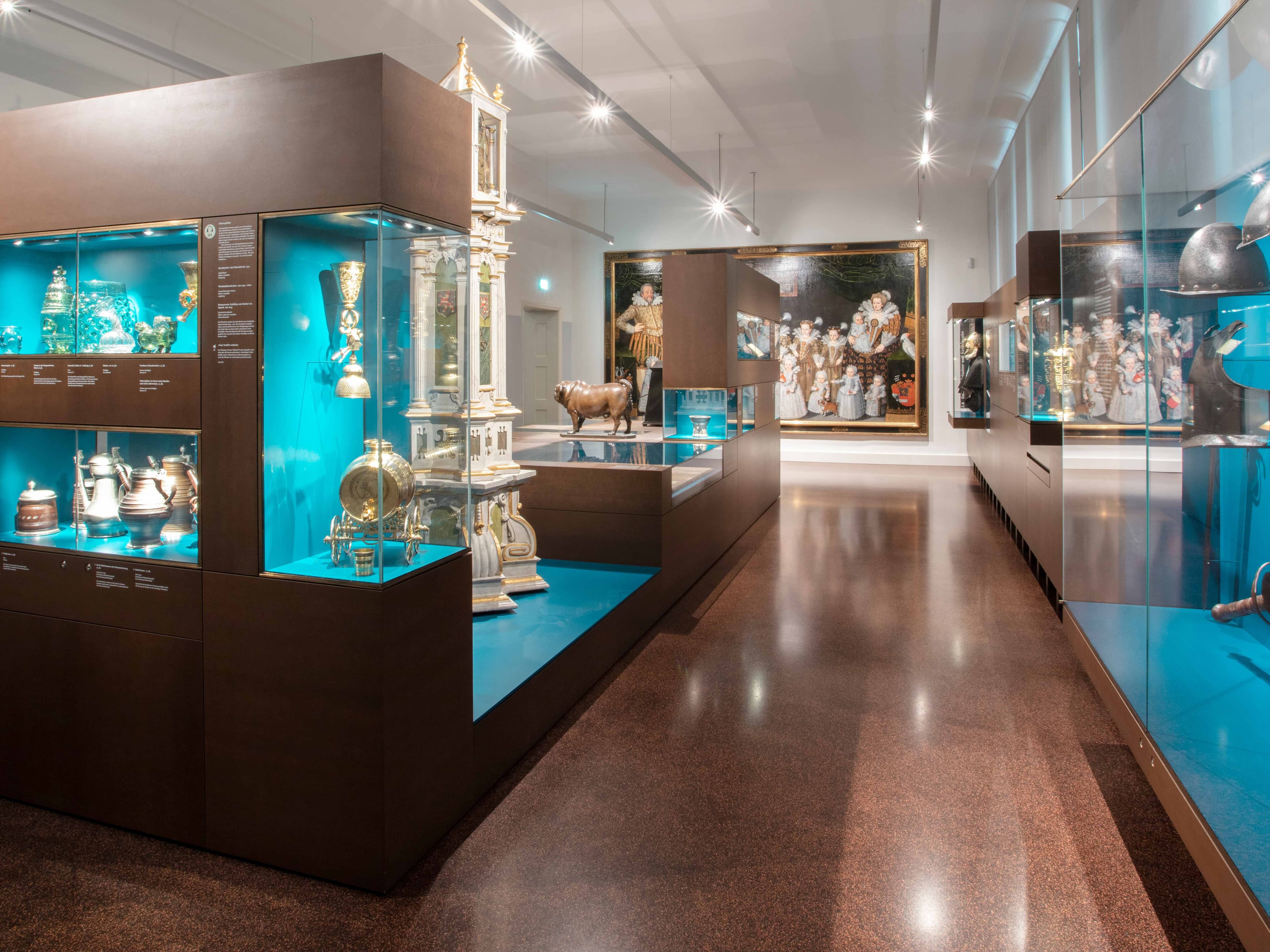 Museumsgalerie mit beleuchteten Vitrinen, in denen Artefakte und ein großes Gemälde an der Wand unter heller Deckenbeleuchtung ausgestellt sind.